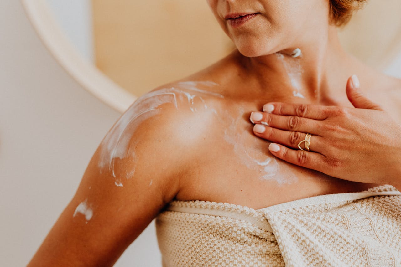 our-story-01 Close-up of a woman applying moisturizing cream to her shoulder indoors, promoting skincare routine.
