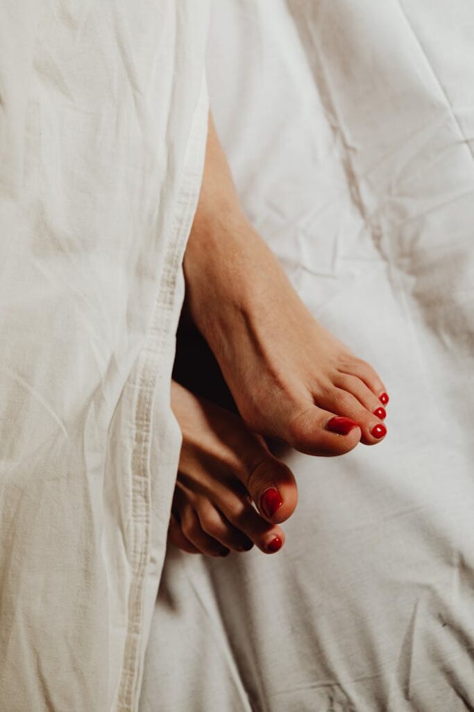 pexels photo 6642887 Close-up of feet with red pedicure peeking out from under white sheets, evoking a relaxed and cozy morning vibe.